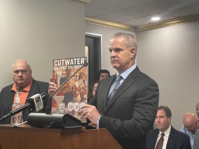 Tom Morgan, representing a spirits distributer, holds up a traditional beer-maker’s advertisement boasting a product’s “real spirits” as he speaks against a controversial wholesaling bill on Tuesday, Jan. 9, 2024. (Leslie Bonilla Muñiz/Indiana Capital Chronicle)