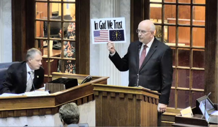 State Sen. Dennis Kruse, R-Auburn, holds a poster Wednesday featuring the phrase "In God We Trust" that he wants all Indiana public schools required to display in every classroom and library. The Senate Education Committee could decide Jan. 15 whether to advance Kruse's Senate Bill 131. iga.in.gov screenshot