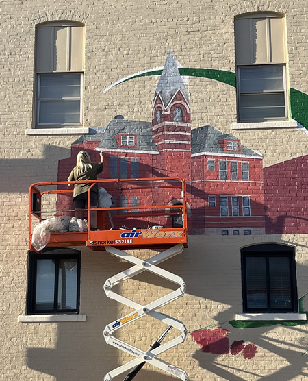 Artist Pam Bliss works on the new mural on the Commerce Street side of the Dunkirk City Building on Monday evening. The public art piece will feature two former Dunkirk high schools, the former Sutton?School and the Dunkirk Speedcats logo. It is expected to be complete by the end of the week, weather permitting. (The Commercial Review/Ray Cooney)