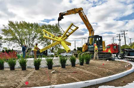 An excavator lifts the sculpture “Bright Days” onto the platform inside the new roundabout at Smith Valley and Averitt roads on Wednesday in Greenwood. The piece had been in place on the Polk Hill Trail as part of Greenwood’s Art on the Trail program since 2019, but will serve as the centerpiece for the new roundabout. RYAN TRARES | DAILY JOURNAL